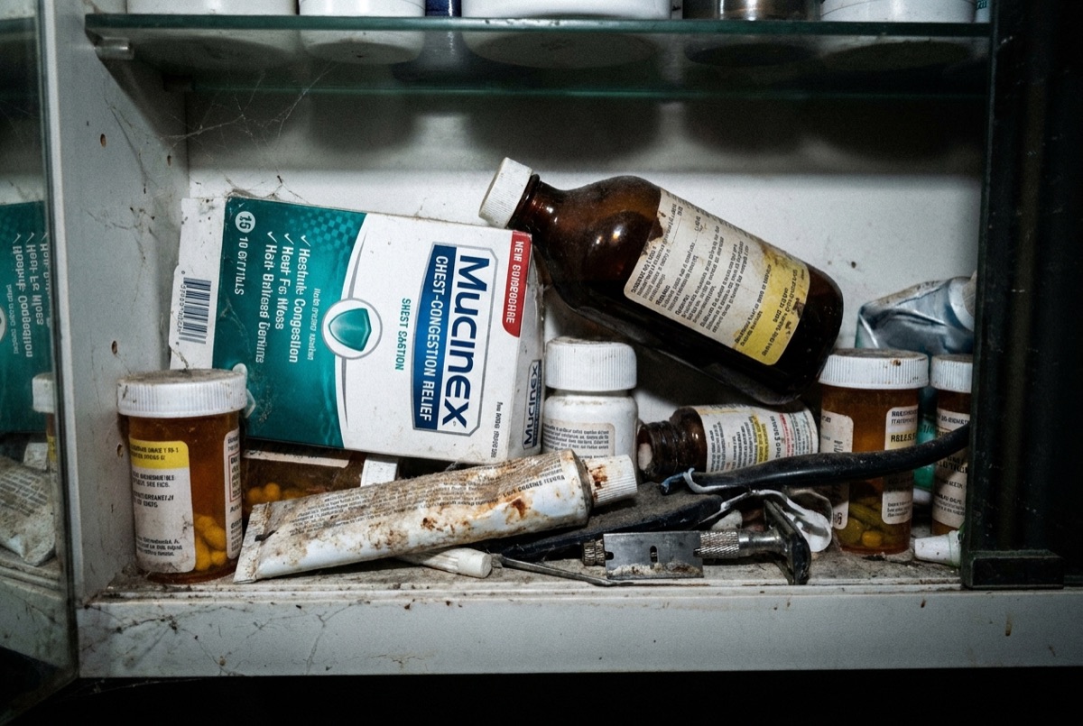 A grimy, dusty medicine cabinet shelf holding a real Mucinex chest-congestion box wedged among old amber pill bottles, a rusty razor and a squeezed-out tube.