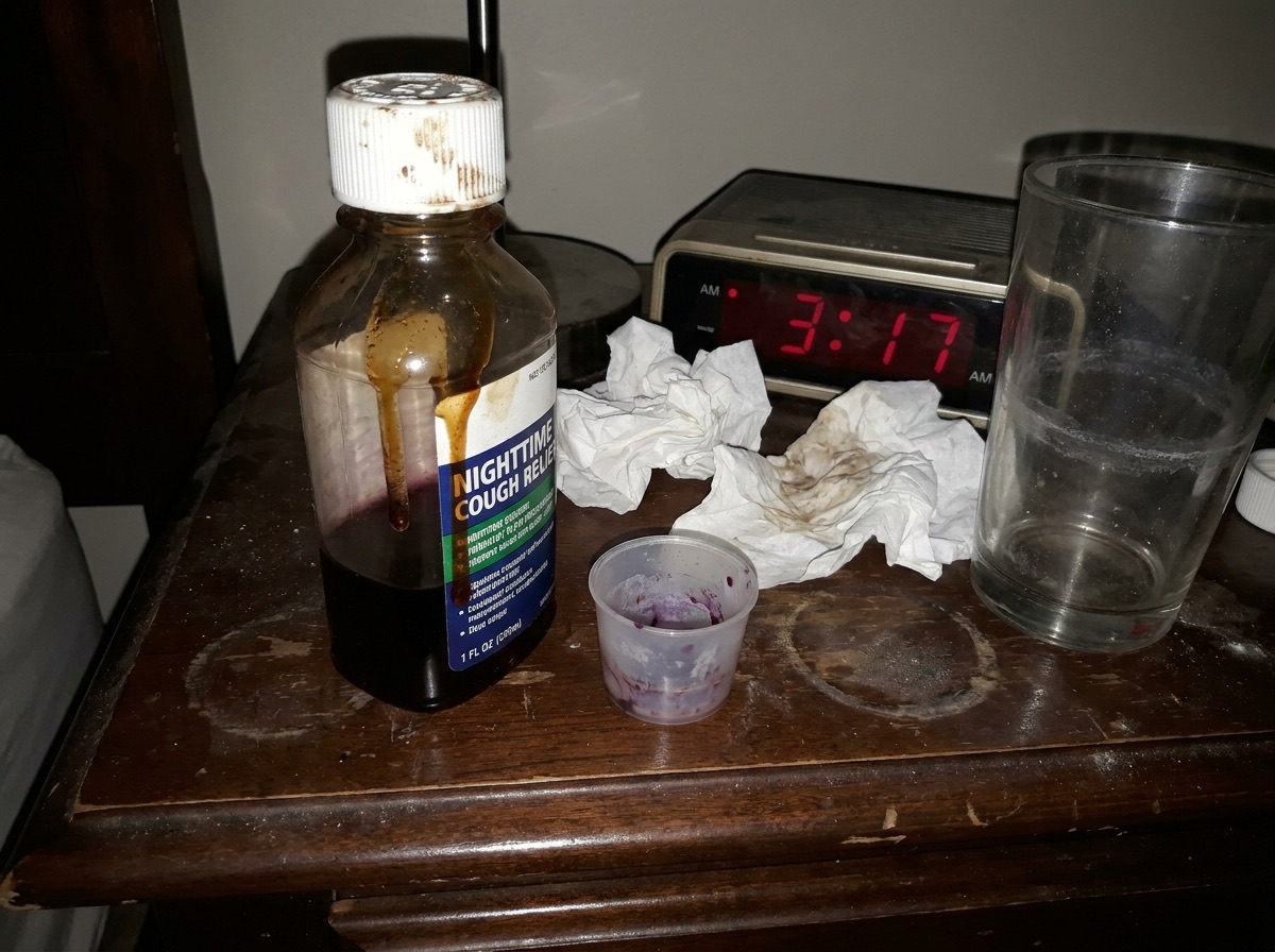 A harsh-flash phone snapshot of a cluttered nightstand at 3 in the morning: a sticky nighttime cough syrup bottle dripping down the side, a stained dosing cup, crumpled tissues and an alarm clock.