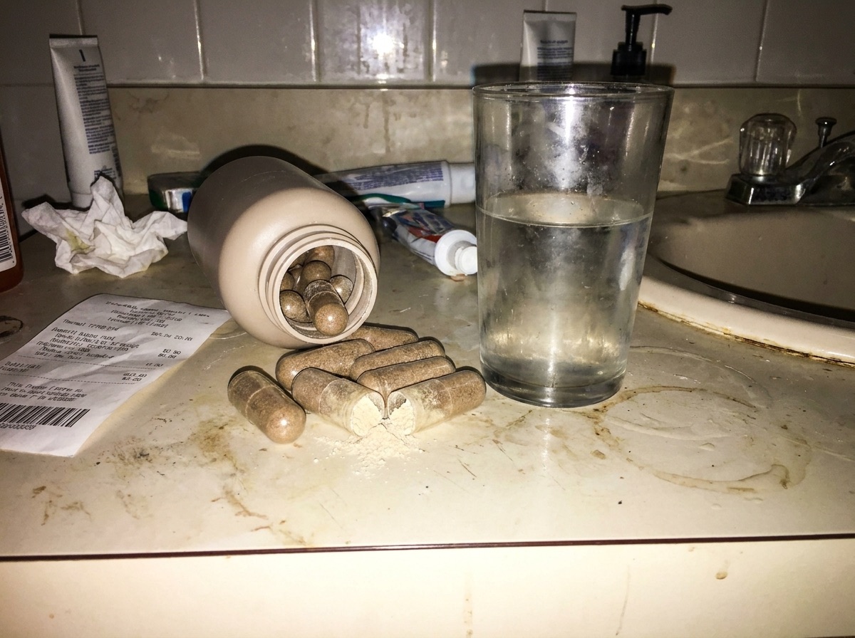 A harsh-flash snapshot of a grimy bathroom counter with a plain bottle tipped over, huge horse-pill capsules spilled out, one cracked and leaking powder, next to a cloudy glass of water.