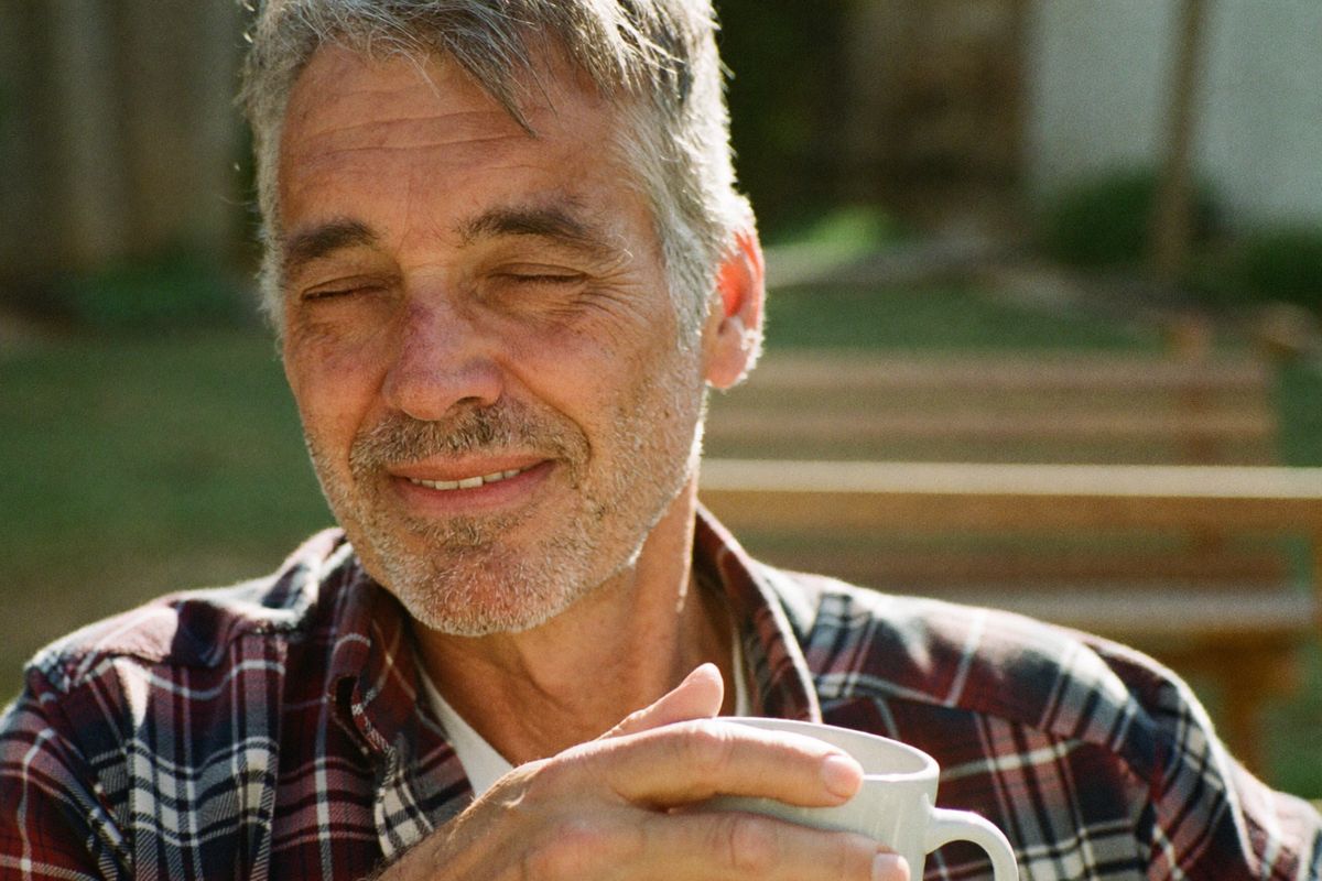 A real, ordinary man in his late fifties on a backyard bench on a bright morning, holding a coffee mug and taking an easy, calm breath.