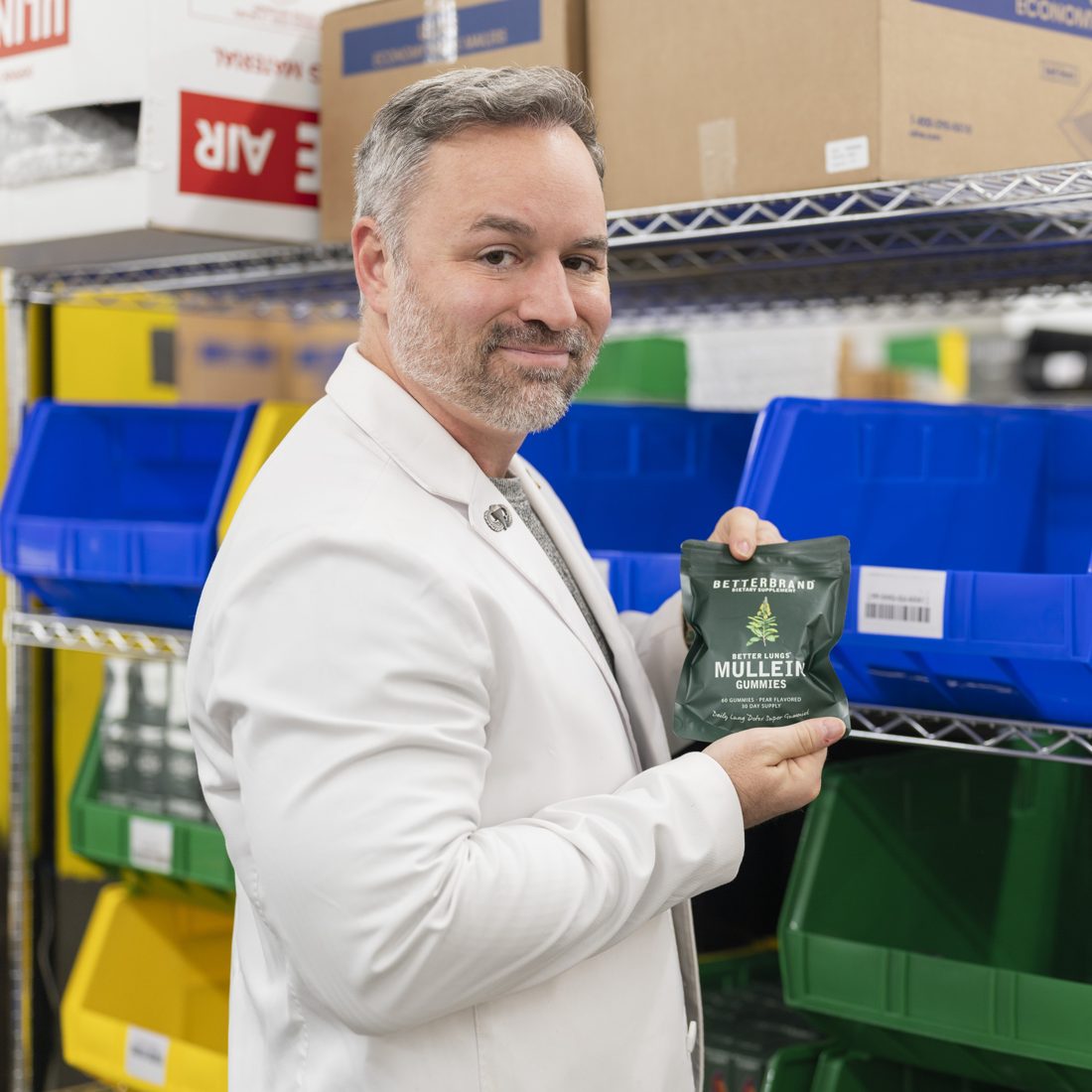 Dr. Chris, PharmD, the pharmacist who founded BetterBrand, in a white coat holding the BetterLungs Mullein Gummies in the fulfillment warehouse.