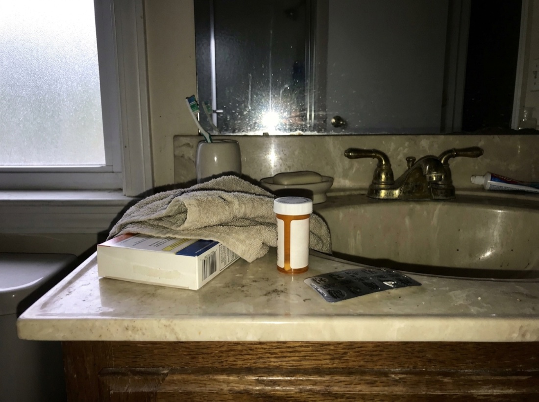 A dated bathroom counter with an over-the-counter chest-congestion box and pill bottle, brand names covered by a folded towel and turned away.