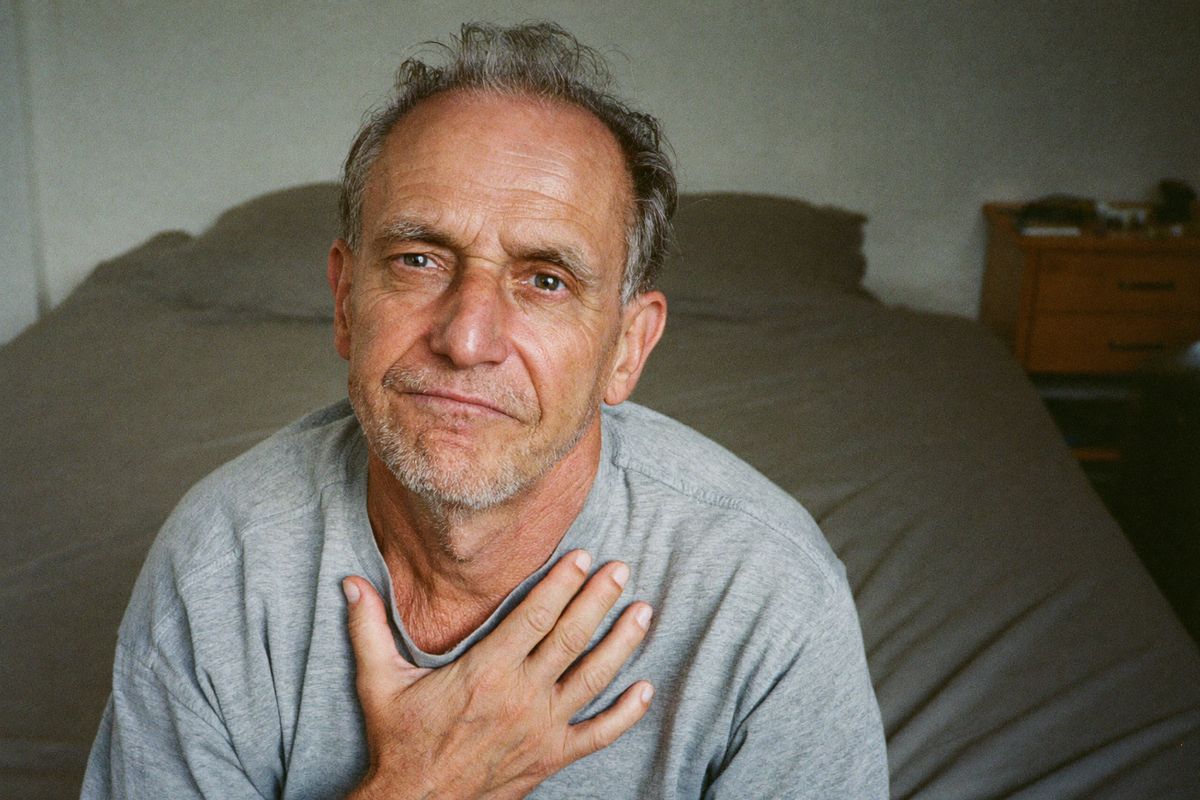 A man in his late fifties sitting on the edge of his bed first thing in the morning, one hand on his chest, clearing his throat.