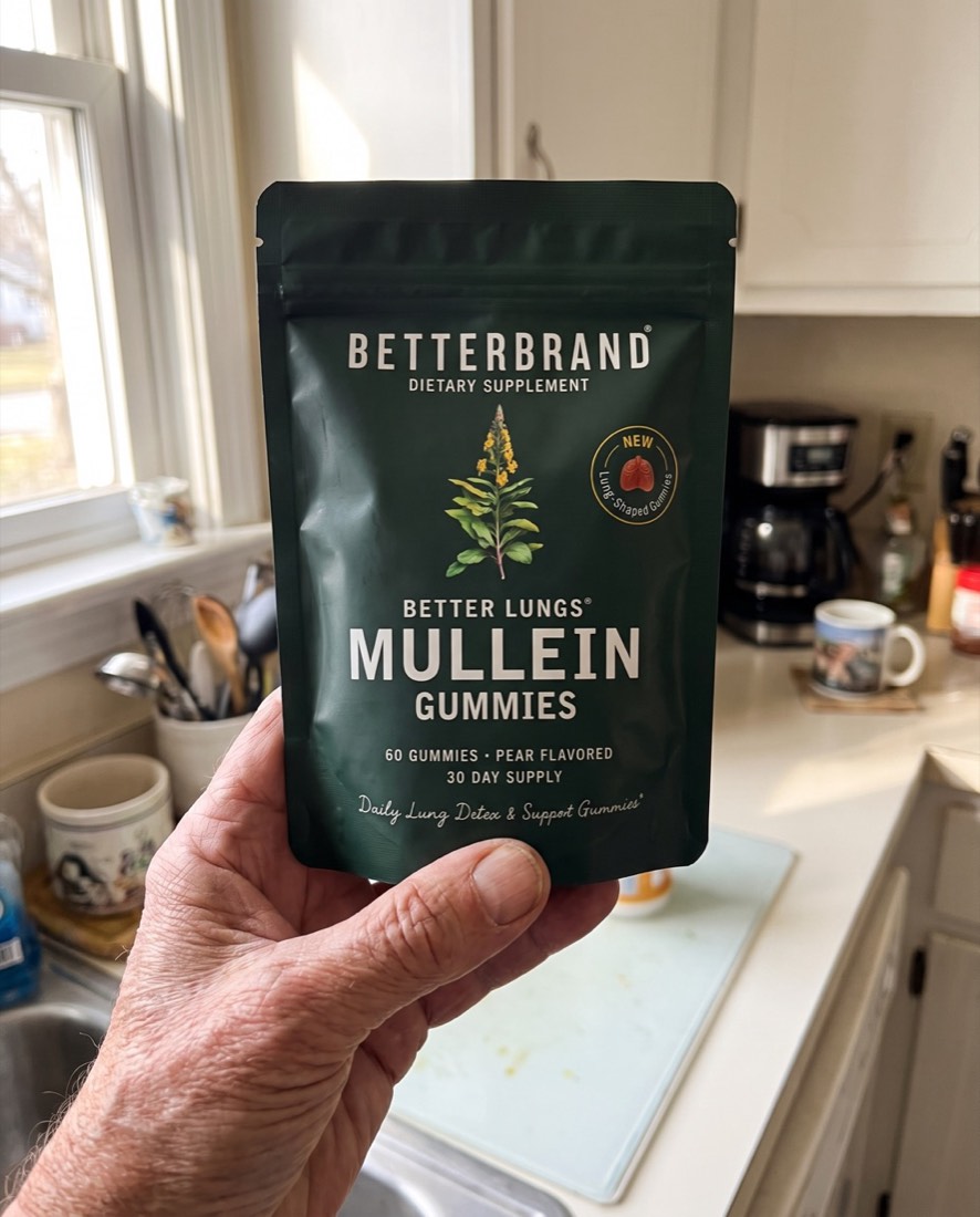 A weathered hand holding up the BetterLungs Mullein Gummies pouch, dark green label facing forward, in a lived-in sunlit kitchen.