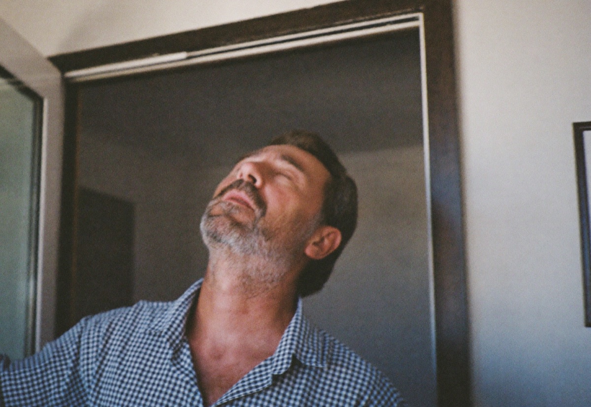A man in his late fifties at an open back door in the morning, eyes closed, taking an easy, relaxed breath.