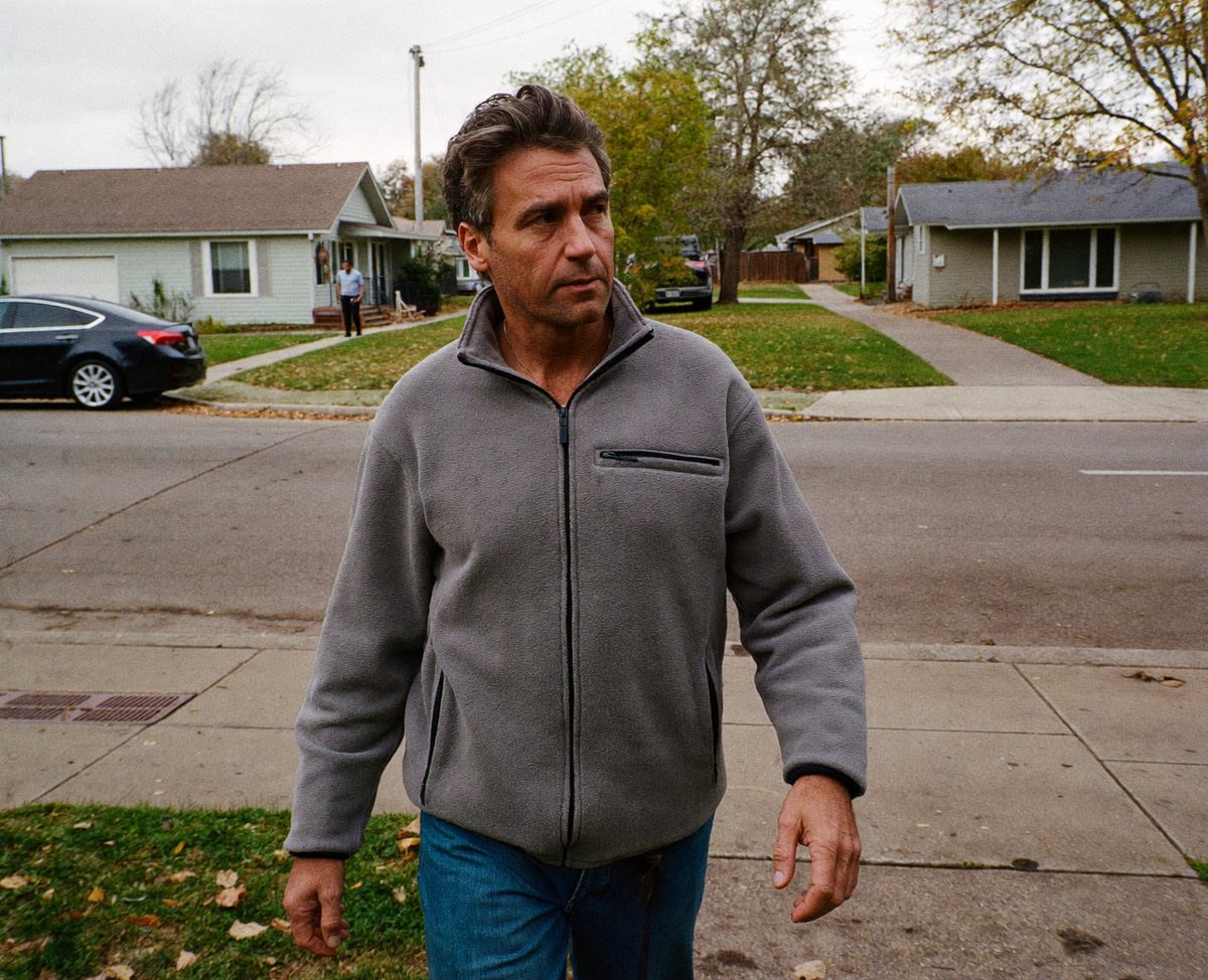A man in his late fifties walking on a neighborhood sidewalk on a regular day, relaxed and unhurried.