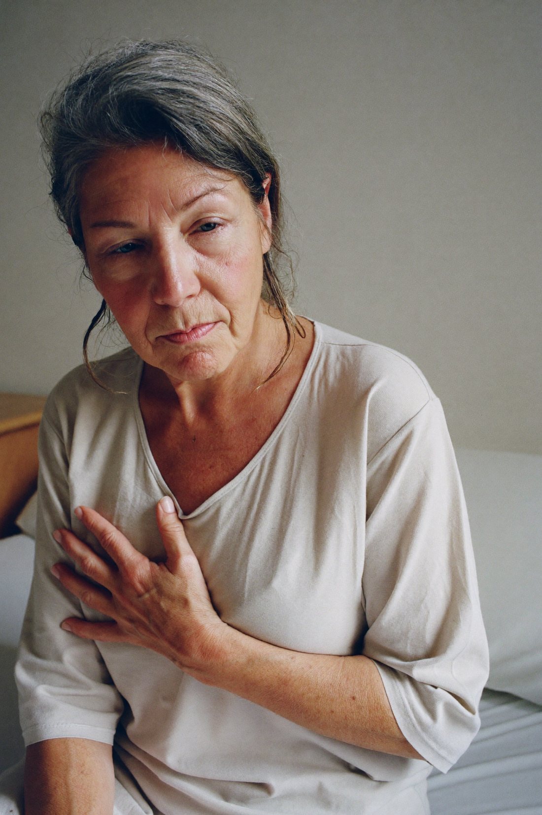 A woman in her late fifties sitting on the edge of her bed in early morning light, clearing her throat with one hand at her chest.