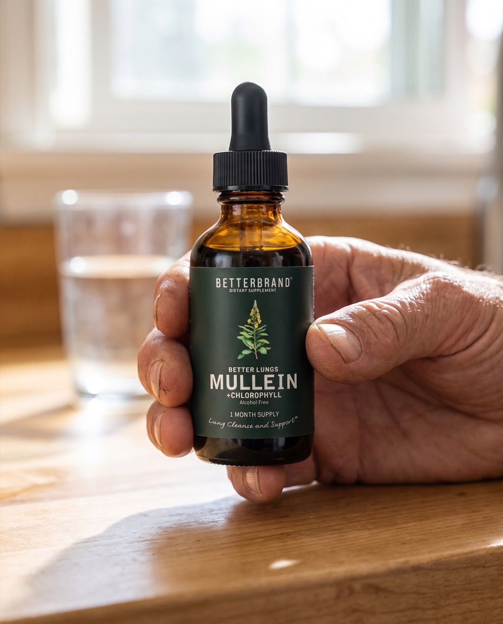 A hand holding the BetterLungs Mullein and Chlorophyll dropper bottle with its dark forest green label, over a glass of water on a sunlit kitchen counter.