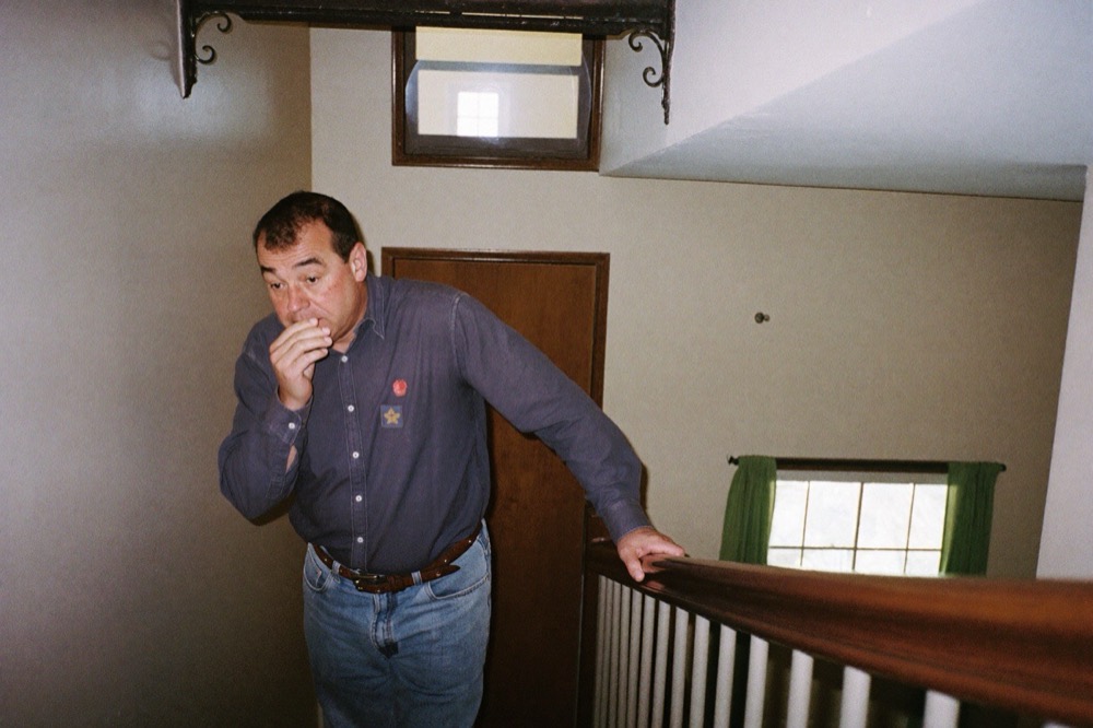 A man in his late fifties pausing halfway up a staircase, one hand on the railing, catching his breath.