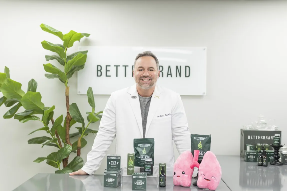 Dr. Chris, a Doctor of Pharmacy in a white lab coat, standing behind a table with the BetterBrand BetterLungs product line and a BetterBrand sign behind him.