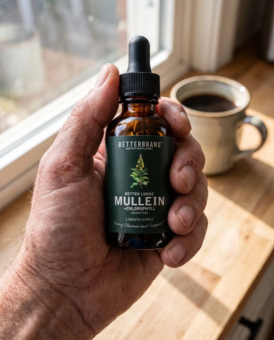 A weathered hand holding the dark forest green BetterLungs Mullein and Chlorophyll dropper bottle, its label facing forward, on a sunlit wooden kitchen counter beside a coffee mug.