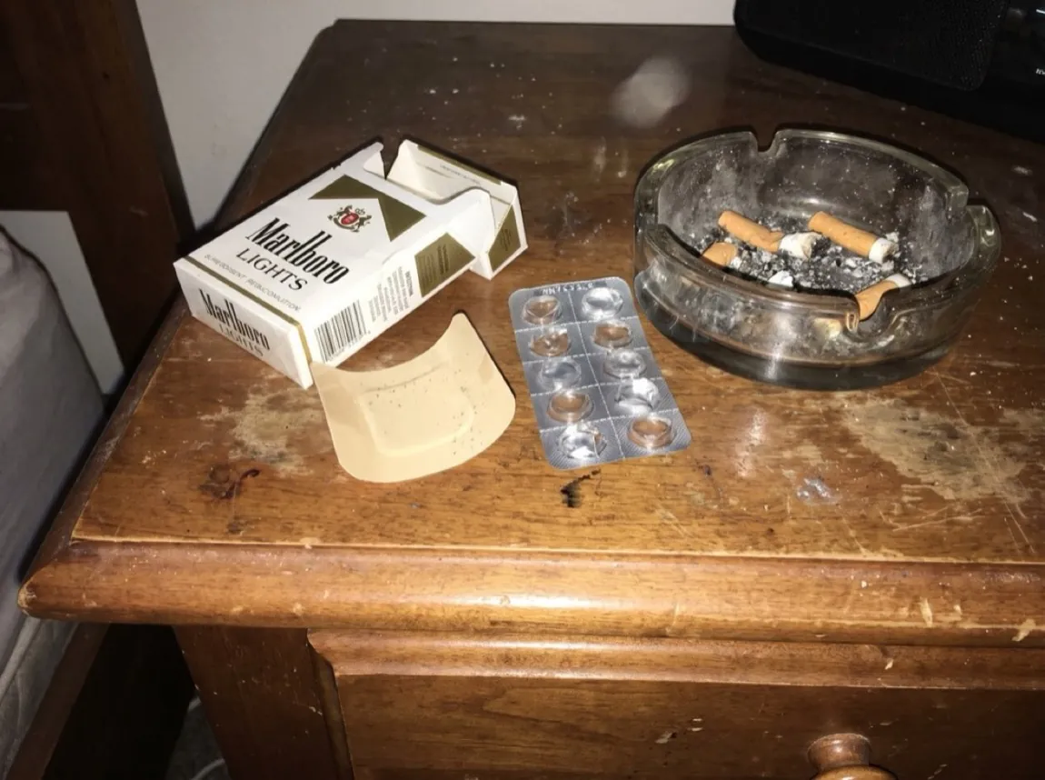 A crushed cigarette pack, a peeled-off used nicotine patch and an empty gum blister on a cluttered nightstand beside an ashtray under a harsh phone flash.