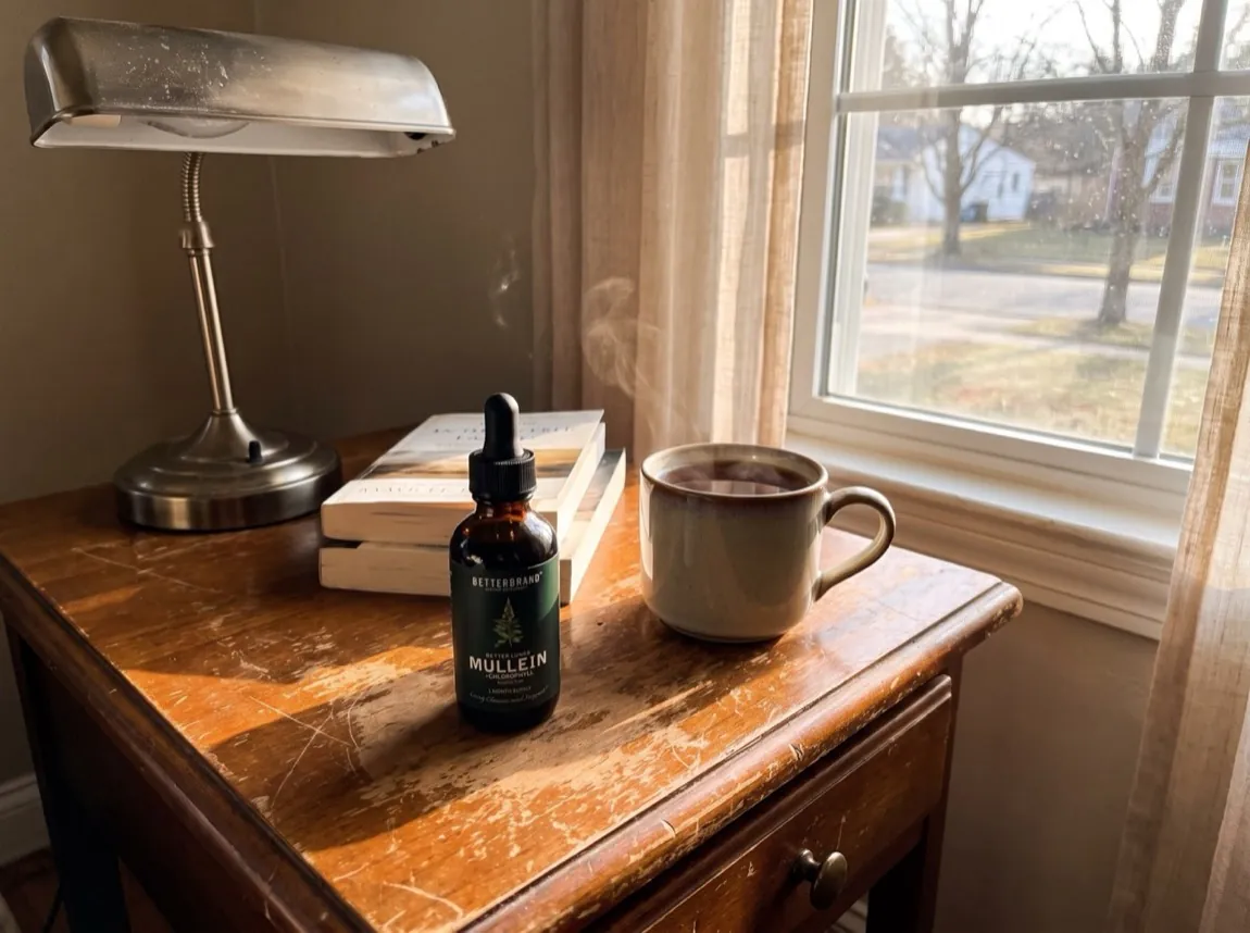 The dark green BetterLungs Mullein and Chlorophyll bottle on a worn wooden side table beside a stack of books and a steaming mug by a sunlit window on a quiet morning.