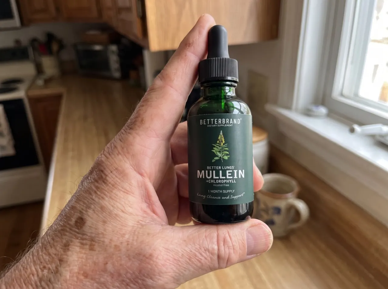 The weathered hands of a woman about 60 holding the dark green BetterLungs dropper bottle and reading the label in natural kitchen light.