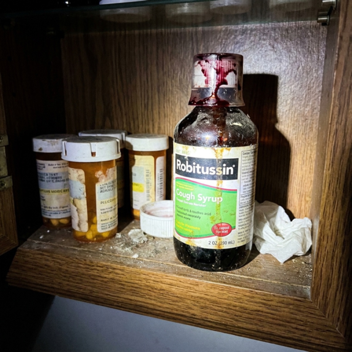A sticky dusty bottle of drugstore cough syrup on a grimy cabinet shelf.