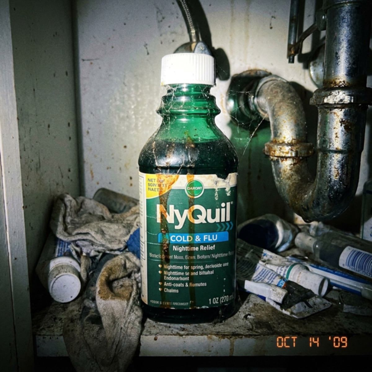 A grimy bottle of nighttime cold and flu liquid shoved under a filthy sink.